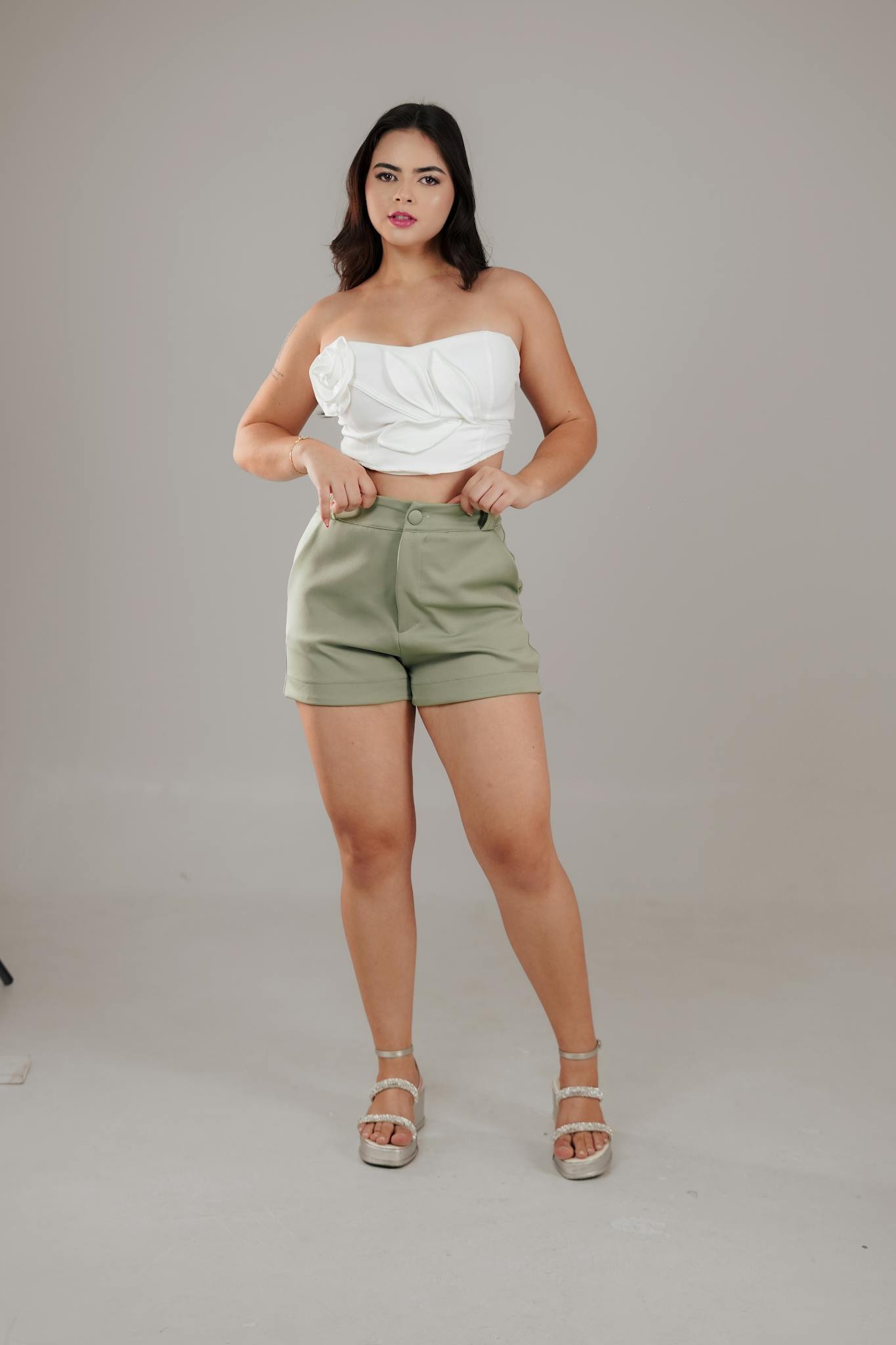 Confident woman in white top and green shorts posing indoors with a neutral background.